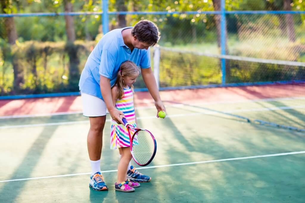 Father and daughter playing tennis on outdoor court. Family with tennis racket and ball at net in sport club. Coach teaching young kid. Child learning to play. Training for kids. Children exercise.